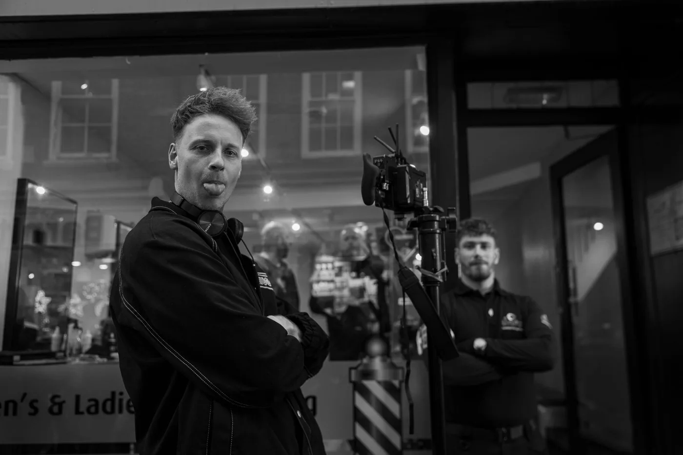 Black and white photograph of a film crew outside a shop at night, a camera on a stand in the foreground and a barber's pole in the window