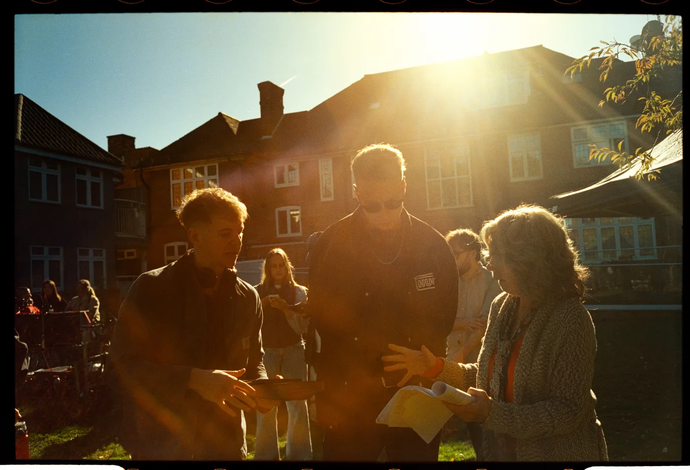 A group talks over a script on a lawn in low autumn sunshine, with a large brick building behind them